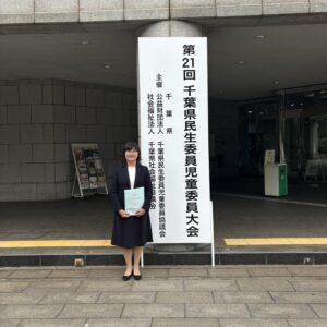 千葉県民生委員児童委員大会の会場入口看板前で入江が資料を手に立つ姿を収めた写真