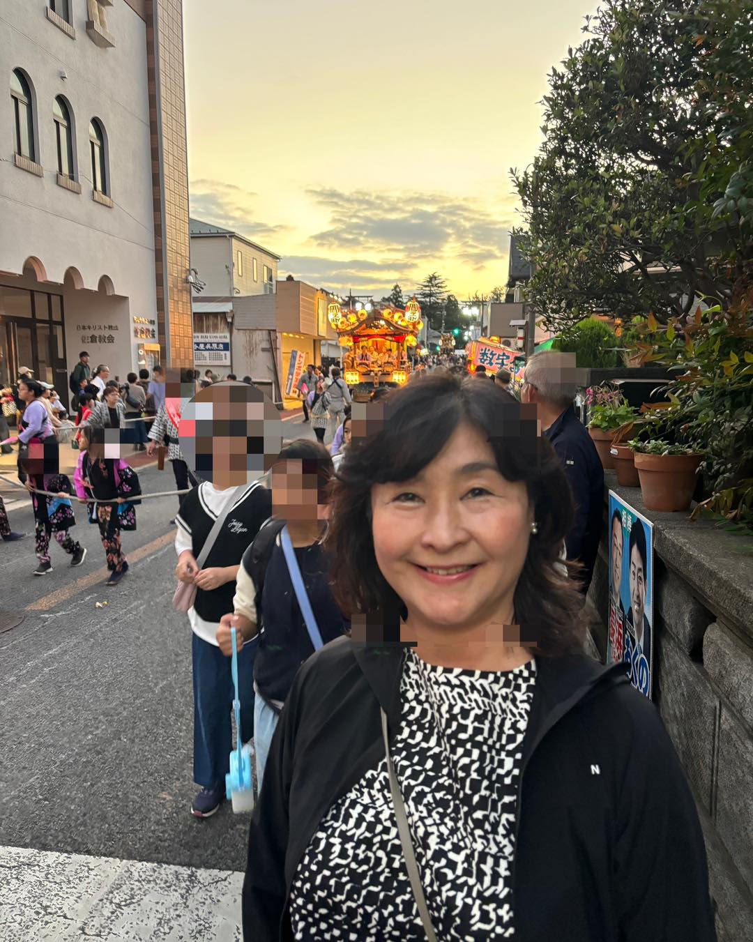 夕暮れの祭り会場で笑顔の入江あき子、背後に山車と人出